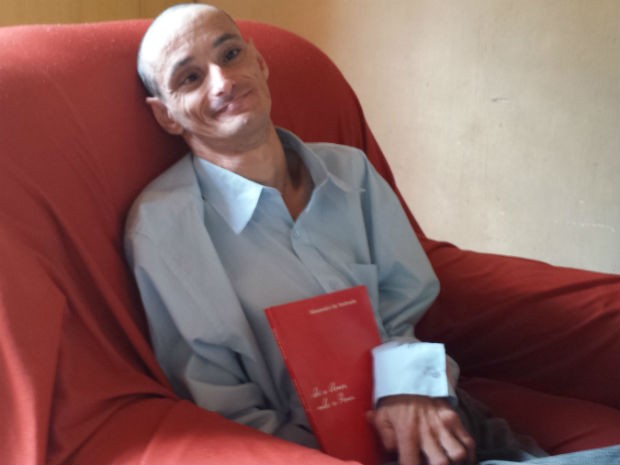 Imagemde Alexandro, vestindo camisa social azul, sentado em uma poltrona vermelha, sorridente, segurando o livro de sua autoria. (Foto: Alana Fonseca/G1). Imagemde Alexandro, vestindo camisa social azul, sentado em uma poltrona vermelha, sorridente, segurando o livro de sua autoria. (Foto: Alana Fonseca/G1).