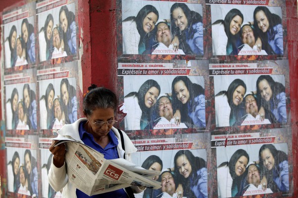 Uma mulher lê o jornal diante de uma parede coberta com cartazes de Hugo Chávez e as filhas em frente ao Hospital Militar de Caracas (Foto: AP Photo/Ariana Cubillos)