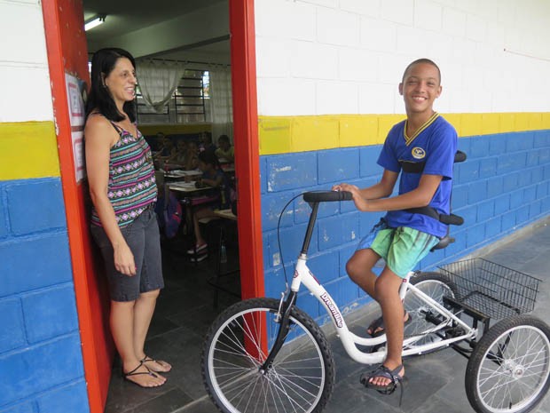 Raphael com a bicicleta ao lado da professora e da sala de aula (Foto: Mariane Rossi/G1)