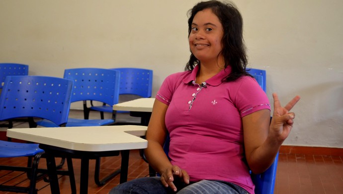 Imagem de Raysa, sentada em sala de aula, sorridente, vestindo uma blusa rosa e calça jeans, fazendo o sinal de paz e amor com a mão esquerda. Foto: Caio Fulgêncio/G1 Imagem de Raysa, sentada em sala de aula, sorridente, vestindo uma blusa rosa e calça jeans, fazendo o sinal de paz e amor com a mão esquerda. Foto: Caio Fulgêncio/G1