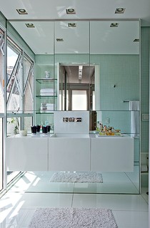 Ainda no projeto de Simone Mantovani, o banheiro de tons claros e com parede de espelhos dá a sensação de amplitude. Repare que a bancada suspensa com gavetões otimiza o espaço