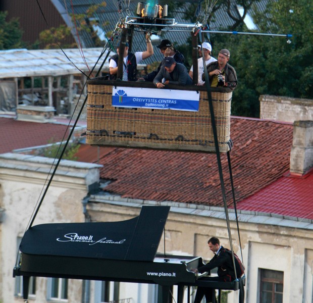 Homem finge tocar piano no ar, enquanto ele e seu instrumento de papelão são levantados por um balão,nesta sexta-feira (10), durante o festival de verão Piano.lt. Vilnius continua sendo uma das poucas capitais europeias que permite balões de ar quente para voar sobre a cidade.  (Foto: AFP)