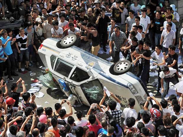 Manifestantes chineses viram carro da polícia em Qidong, durante protesto contra fábrica que pretendia jogar resíduos poluentes e contaminar água na região. (Foto: Kyodo News / AP Photo)