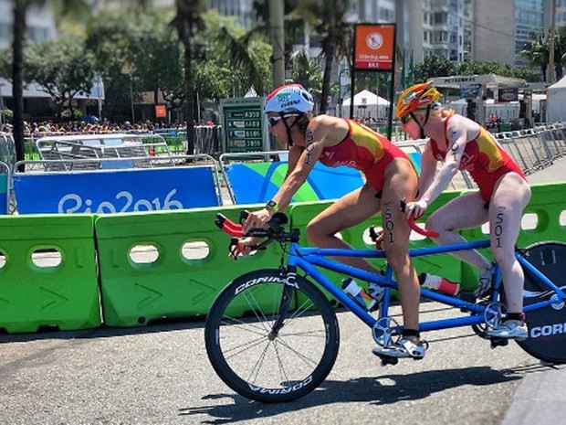 João Maia faz registro do ciclismo na Paralimpíada (Foto: Reprodução / Instagram) Foto feita na prova de triatlo para deficiente visual. Vindo da direita para esquerda, atletas mulheres em uma bicicleta de dois lugares na cor azul, com a roda dianteira raiada e a traseira fechada com tampão corta vento na cor preta. A atleta que guia a bicicleta tem pele bronzeada, veste maiô vermelho e amarelo e usa capacete branco. A atleta sentada atrás tem pele muito branca, veste maiô da mesma cor da companheira e capacete nas cores vermelho e amarelo. Delimitando o traçado do circuito existem divisórias plásticas na cor verde claro e ao fundo estão os prédios da orla de Copacabana.