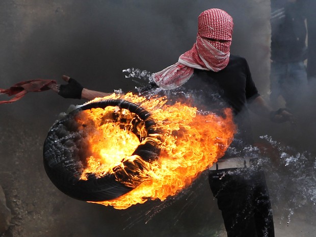 Palestino atira um pneu em chamas contra soldados israelenses durante conflito em Betunia, perto de Ramallah, Cisjordânia. Centenas de manifestantes entraram em choque com militares israelenses após uma marcha marcando os 65 anos do 'Dia da Catástrofe'. (Foto: Abbas Momani/AFP)