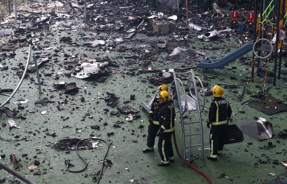 Bombeiros passam por playground coberto de destroços enquanto trabalham no combate ao fogo em uma torre residencial de Londres. na Inglaterra (Foto: Neil Hall/Reuters)