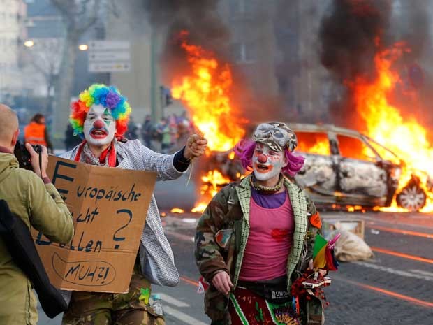 Manifestantes vestidos de palhaços passam por um carro da polícia incendiado. (Foto: Michael Probst / AP Photo)