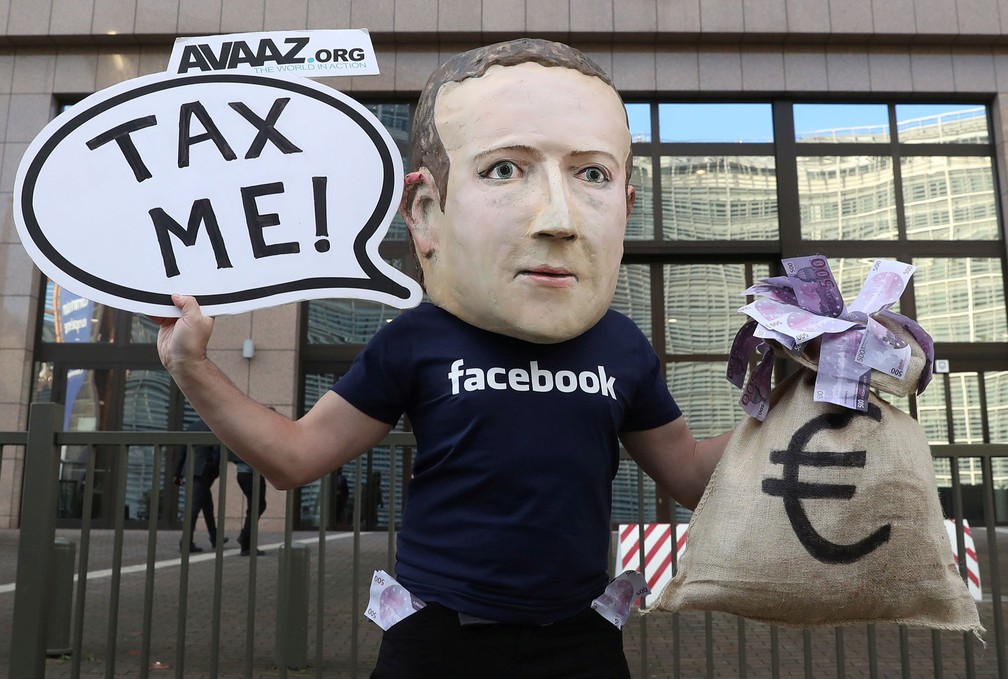 Um ativista protesta usando uma máscara do fundador do Facebook, Mark Zuckerberg, um saco de dinheiro e o cartaz que diz 'Me taxem',  em frente à sede da União Europeia em Bruxelas, na Bélgica, aproveitando a reunião de ministros de Finanças do grupo — Foto: Yves Herman/Reuters