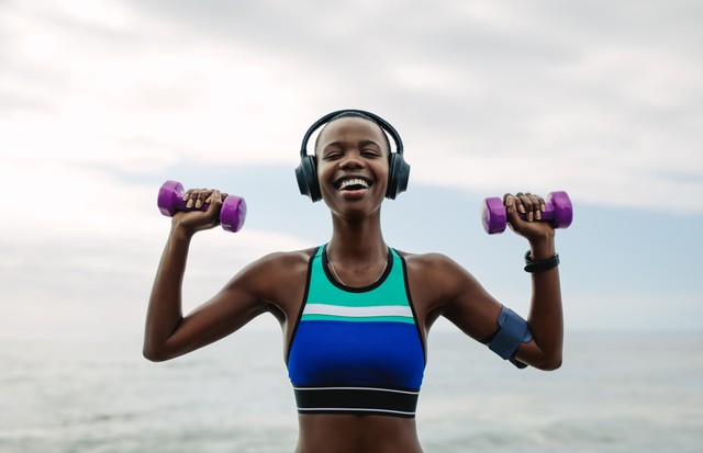Happy woman wearing headphones exercising with dumbbells outdoors. Fitness female wearing headphones doing workout by the beach in morning. (Foto: Getty Images/iStockphoto)