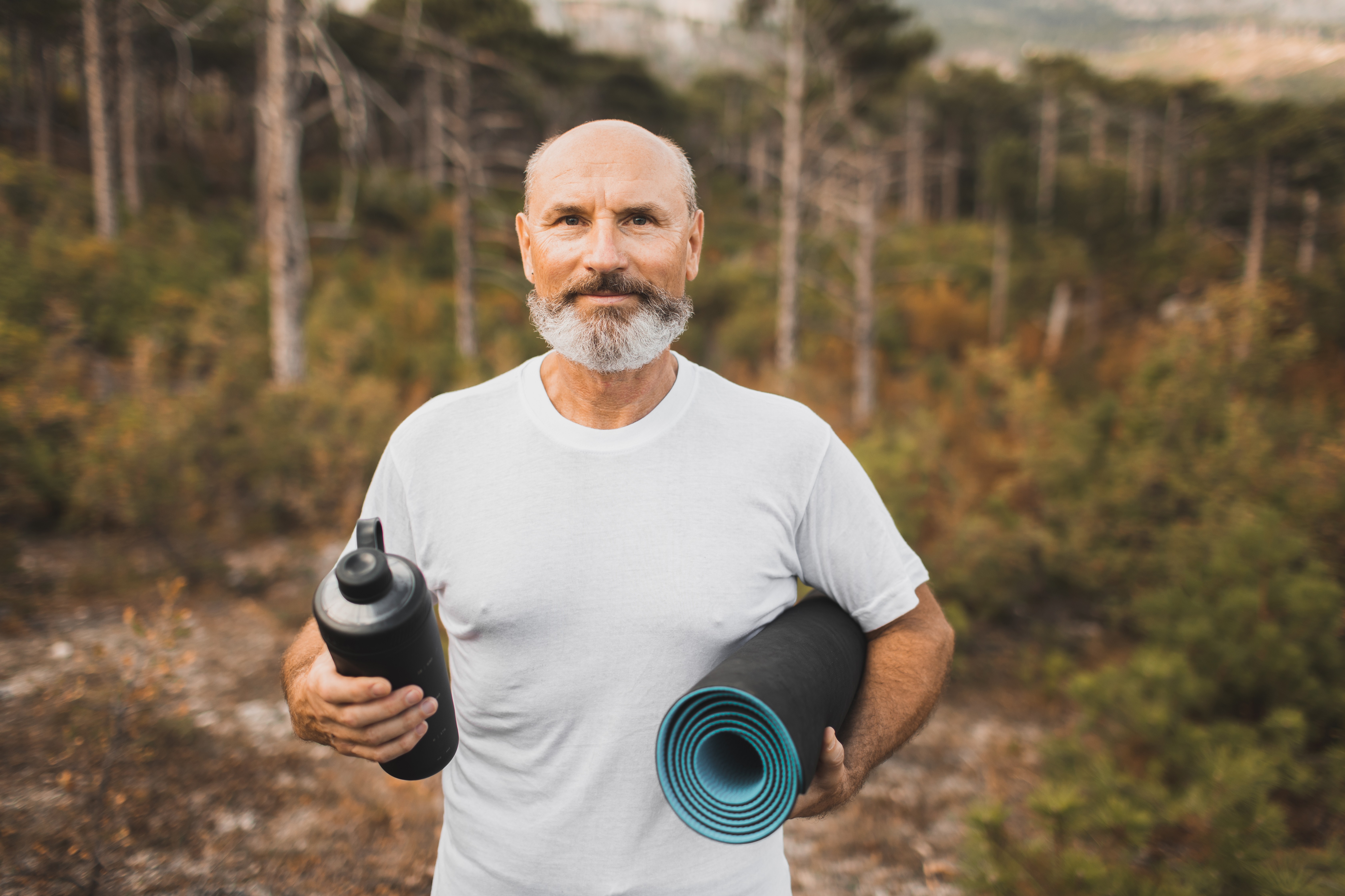 Portrait of senior yoga coach with reusable water bottle and mat in white t-shirt outdoors in autumn forest. Healthy lifestyle in old age. Active old man with gray beard, elderly generation. Yoga guru. FItness and sports training outdoors. Happy on retire (Foto: Getty Images)