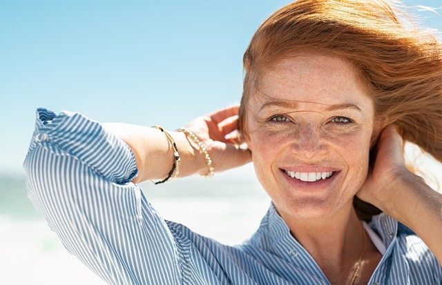 Portrait of beautiful mature woman with wind fluttering hair. Closeup face of healthy young woman with freckles relaxing at beach. Cheerful lady with red hair and blue blouse standing at seaside enjoying breeze looking at camera. (Foto: Getty Images/iStockphoto)
