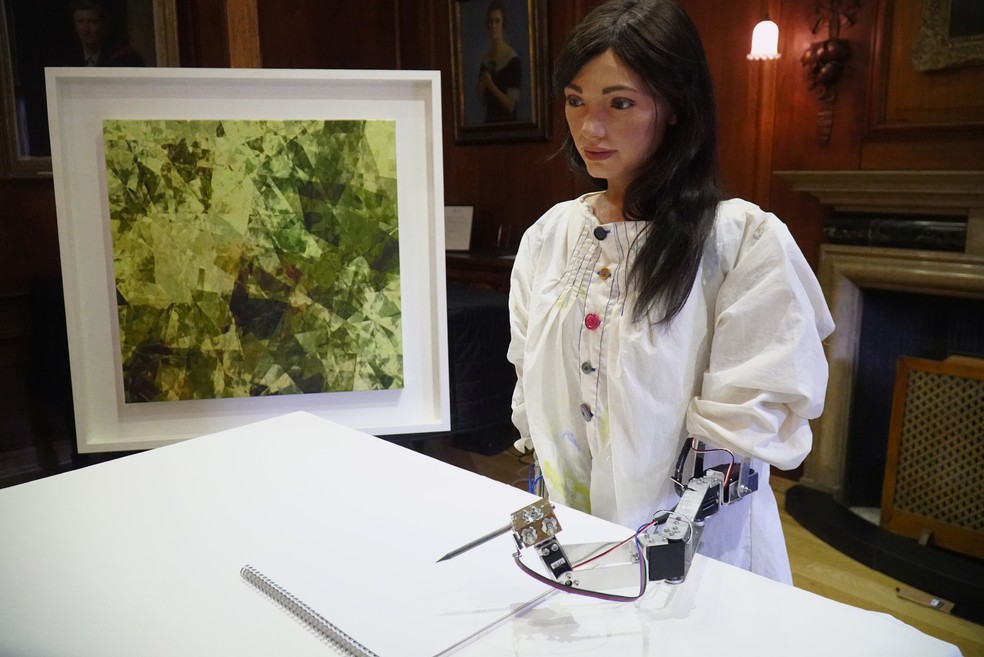 A robô Ai-Da usa um lápis preso a seu braço para fazer um desenho, ao lado de uma pintura baseada em sua visão — Foto: Matthew Stock/Reuters