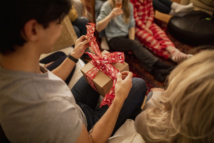 A group of friends are sitting together in the living room at a house party, opening Christmas presents. (Foto: Getty Images)