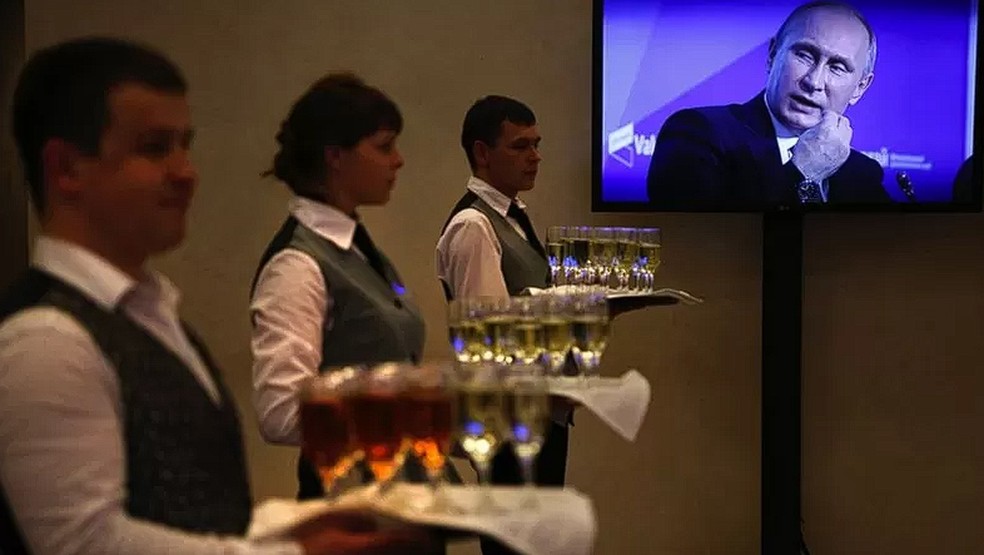 Garçons são vistos servindo bebidas em taças diante da imagem do presidente russo, Vladimir Putin — Foto: GETTY IMAGES/via BBC
