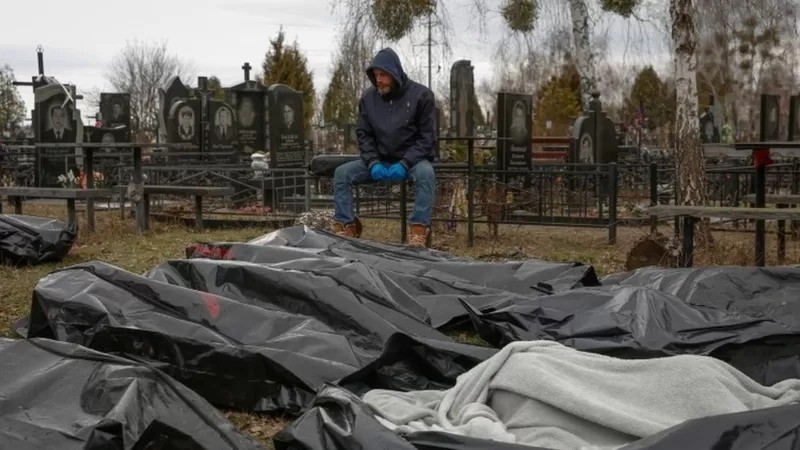 Um funcionário do serviço funerário senta-se ao lado de corpos de civis em Bucha (Foto: Reuters via BBC News )