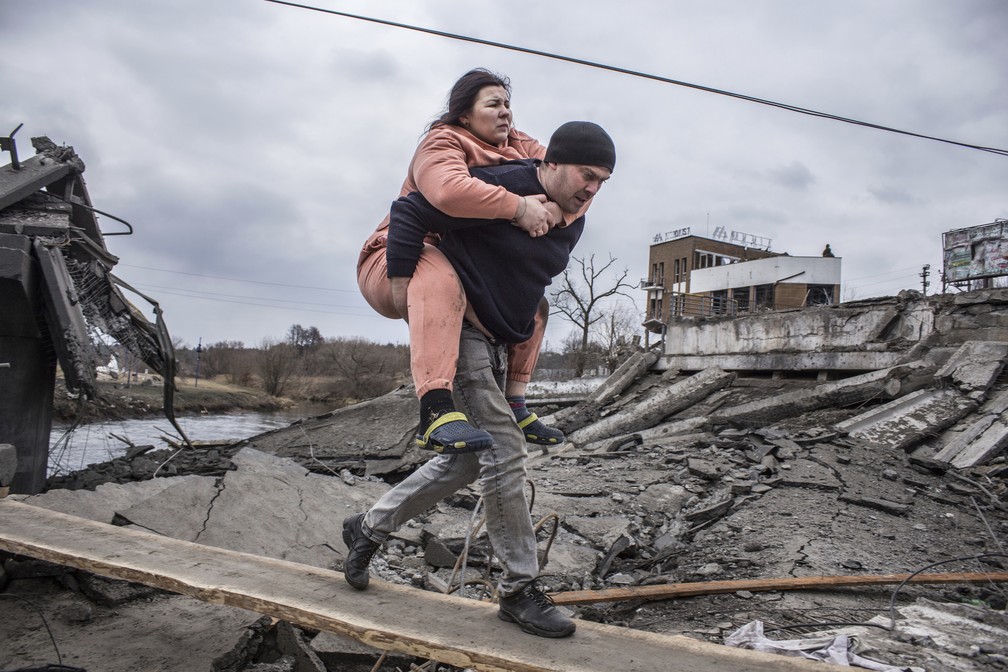 Homem carrega mulher nas costas em caminho improvisado sobre os escombros na cidade de Irpin — Foto: Oleksandr Ratushniak/AP Photo