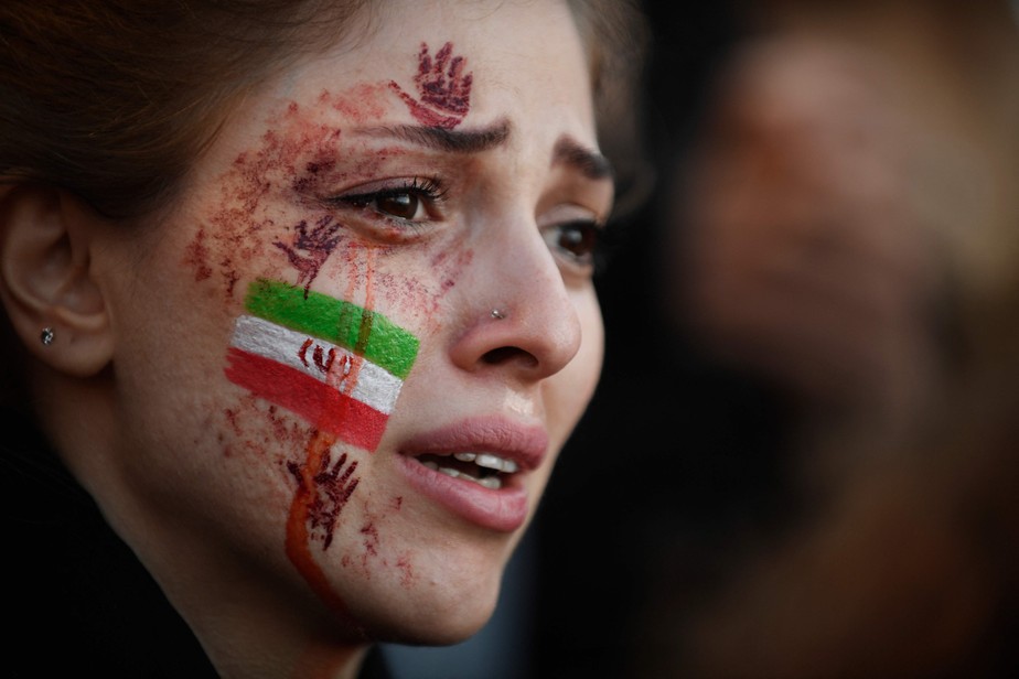 Manifestante com bandeira iraniana e mãos vermelhas pintadas no rosto participa de manifestação em apoio aos protestos iranianos, em Paris