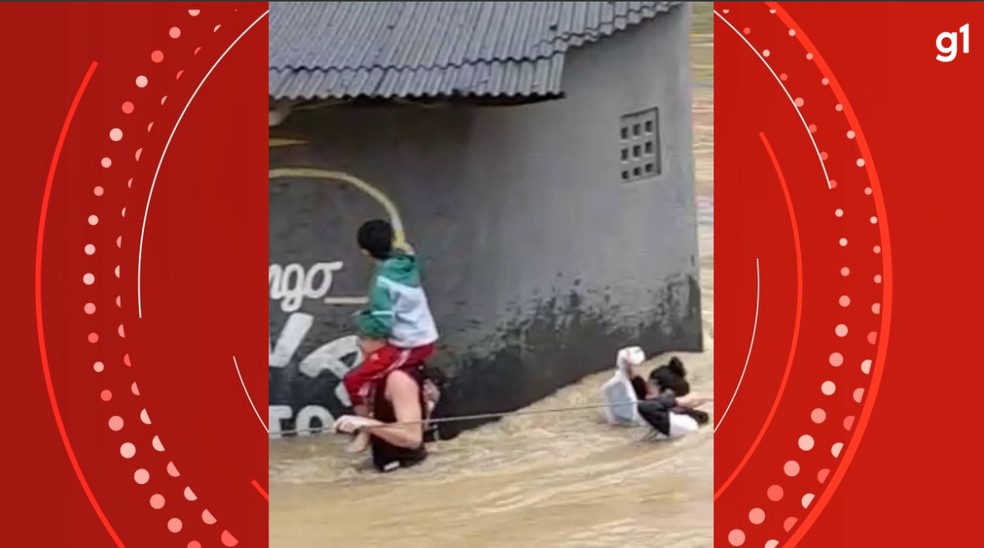 Homem se segura em corda e passa por rua alagada com criança na cabeça — Foto: Reprodução/Redes sociais