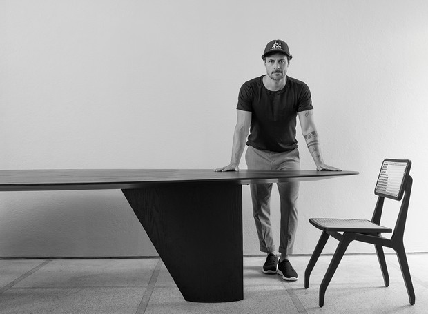 O designer está apoiado na mesa Lago, criada por ele em 2021, e ao lado da cadeira Lenzi, de 2010 (Foto: Daniel Mansur / Editora Globo)