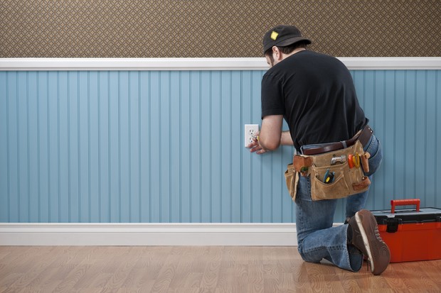 Male handyman with toolbox and tool belt working on power outlet in empty domestic room. The wall has a blue beadboard wainscoting and a patterned wallpaper. (Foto: Getty Images)