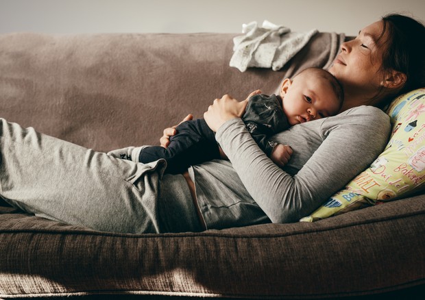 Side view of a woman lying on couch with eyes closed and holding her baby on her chest. Tired mother taking rest sleeping on a sofa with her baby on her. (Foto: Getty Images/iStockphoto)