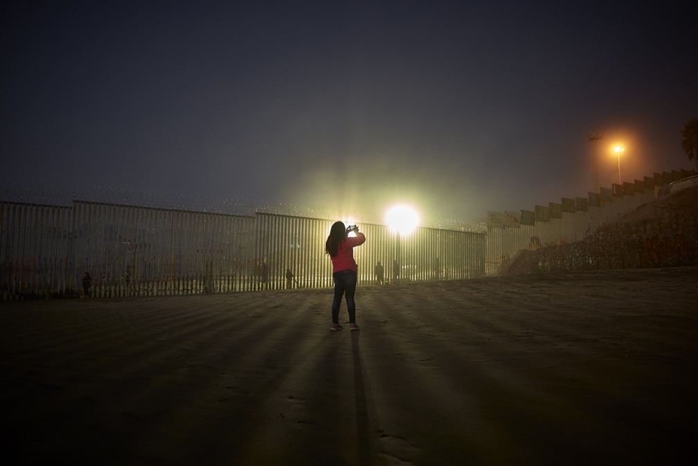Mulher faz imagens com telefone, enquanto holofotes dos Estados Unidos iluminam a parede com arame farpada na fronteira, ao longo da praia em Tijuana, no MÃ©xico â Foto: AP Photo/Gregory Bull