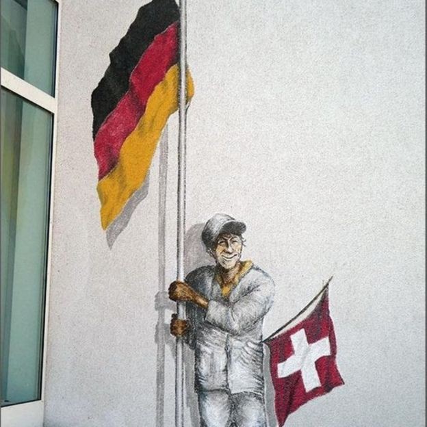 BBC - A Trilha do Enclave (ou Exclave) inclui 11 paradas em Büsingen, como esse mural de um homem segurando uma bandeira alemã e uma suíça (Foto: LARRY BLEIBERG/BBC)