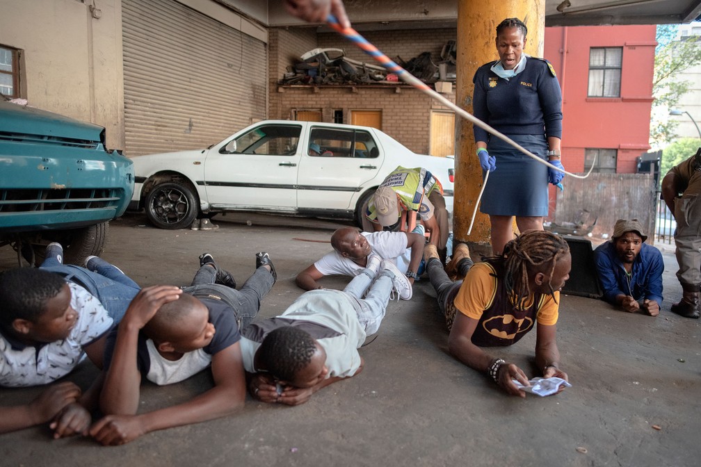 Integrante do Serviço de Polícia da África do Sul faz com que suspeitos de violarem regras de isolamento fiquem deitados no chão em Joanesburgo, na África do Sul, na sexta-feira (27)  — Foto: Luca Sola / AFP