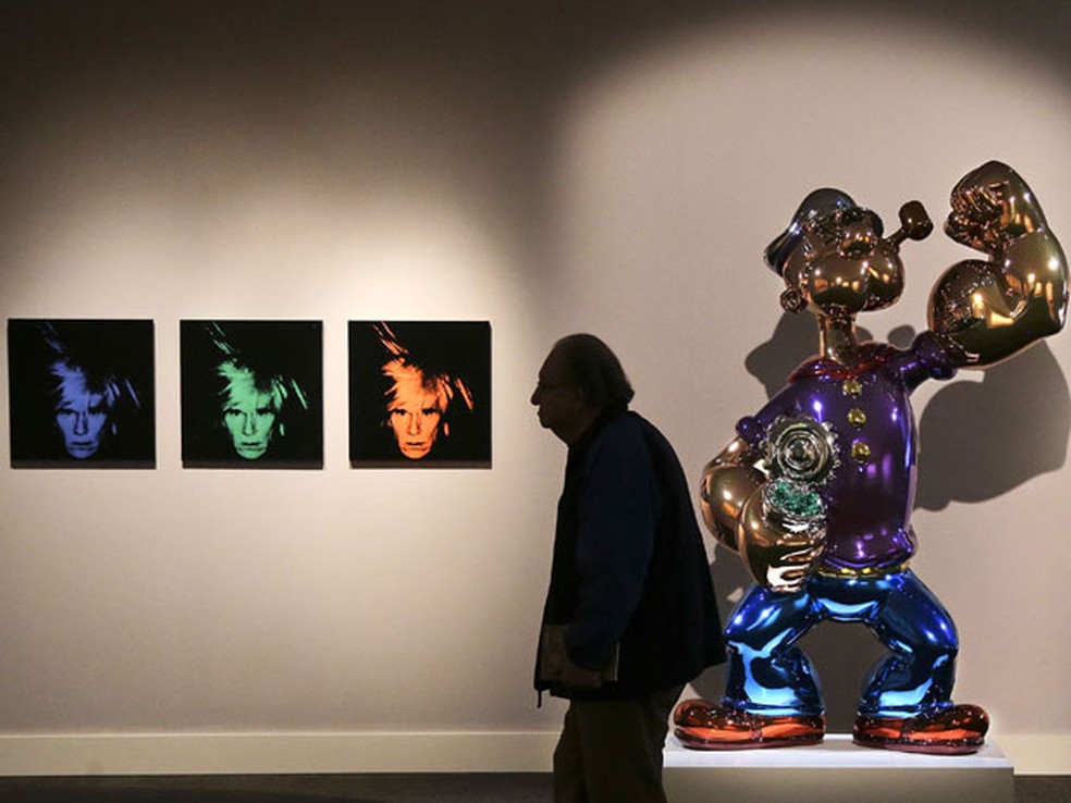 Possível comprador passa diante da obra Six Self Portraits, de Andy Warhol, e Popeye, de Jeff Koons, durante exibição prévia do leilão da Sotheby's, em Nova York — Foto: Julie Jacobson/AP