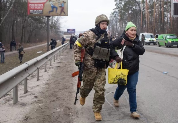 Militar ucraniano ajuda mulher a fugir de região sob ataque russo; símbolo adicionado por ele ao uniforme chamou a atenção nas redes sociais por ser associado ao nazismo (Foto: ANASTASIA VLASOVA/GETTY IMAGES via BBC)