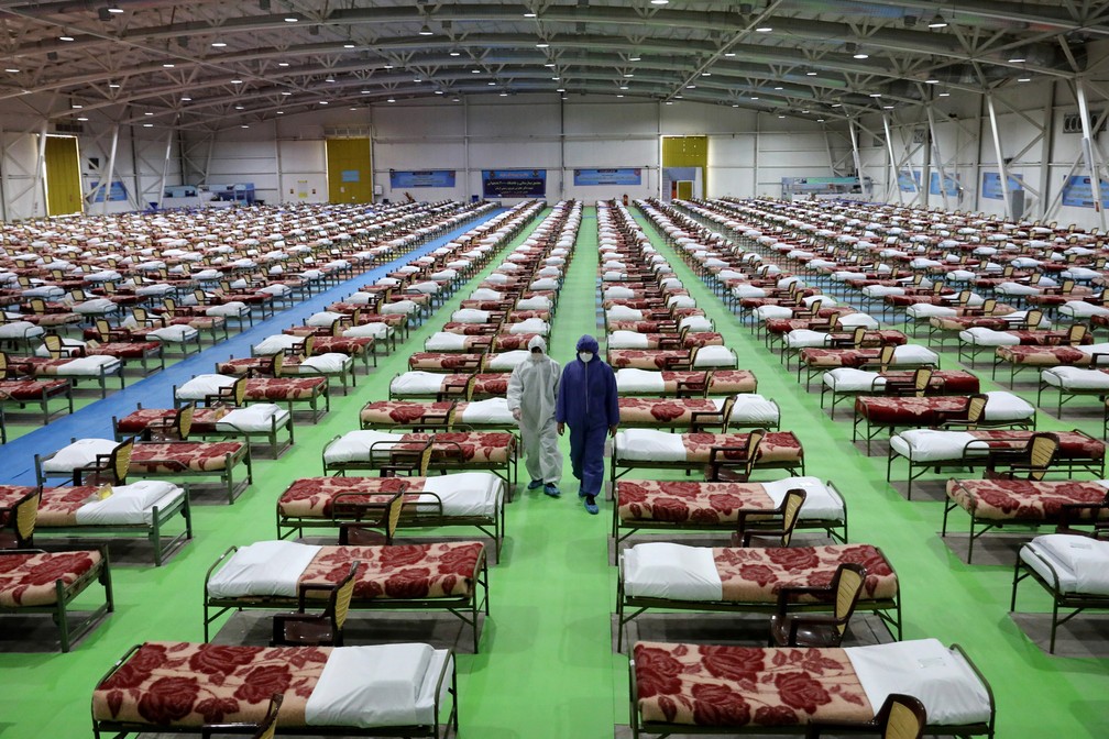 Pessoas com roupas de proteção passam por filas de camas em um hospital temporário de 2000 leitos criado pelo exército iraniano para pacientes com o novo coronavírus no centro de exposições internacional no norte de Teerã, no Irã, em 26 de março — Foto: Ebrahim Noroozi/AP