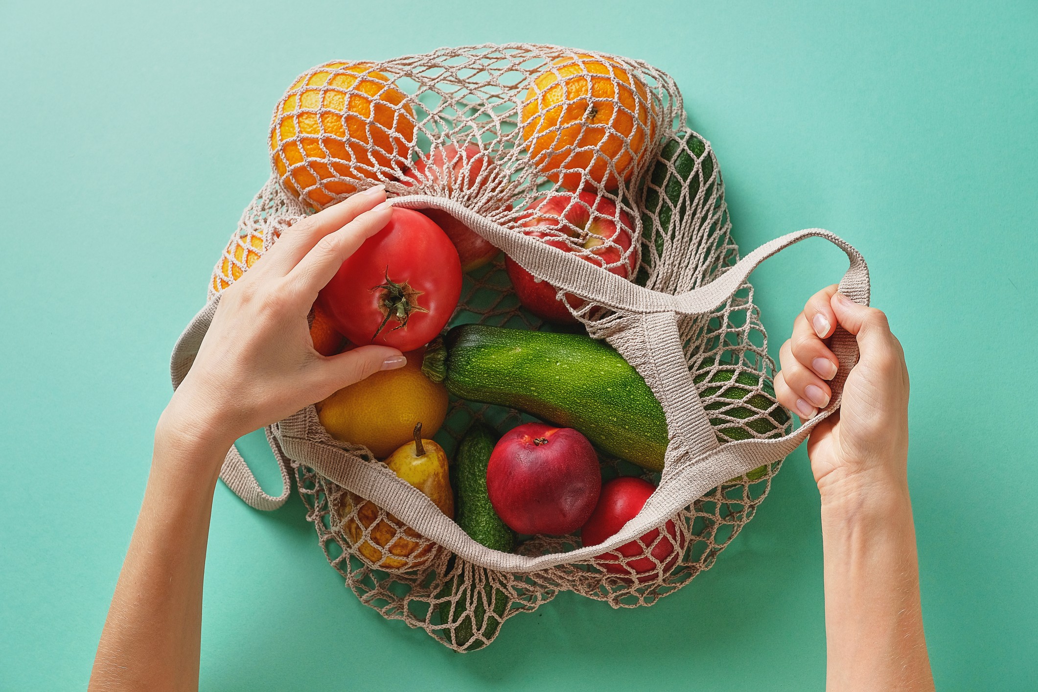 Fresh juicy fruits and vegetables, products in a reusable shopping bag. A girl or woman takes or lays out products from a string bag made from recycled materials on a Green Pastel background. Vegetarianism, Veganism. No plastic. (Foto: Getty Images)