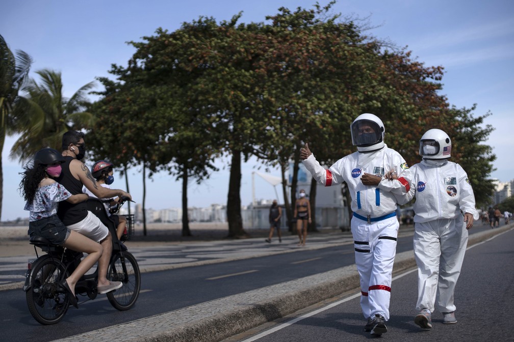 12 de julho: casal anda pela calçada na praia de Copacabana e acena para pessoas em uma moto. O casal veste roupas protetoras contra a Covid-19 que se assemelham a vestes de astronautas. — Foto: Mauro Pimentel/AFP