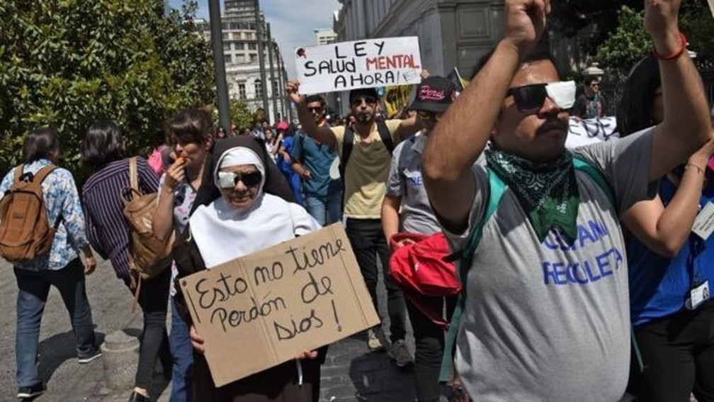 Com olho tampado, mulher vestida de freira ergue cartaz que diz: 'Isso não tem perdão de Deus'; outro manifestante no Chile expõe cartaz: 'Lei para saúde mental agora!' — Foto: Getty Images via BBC