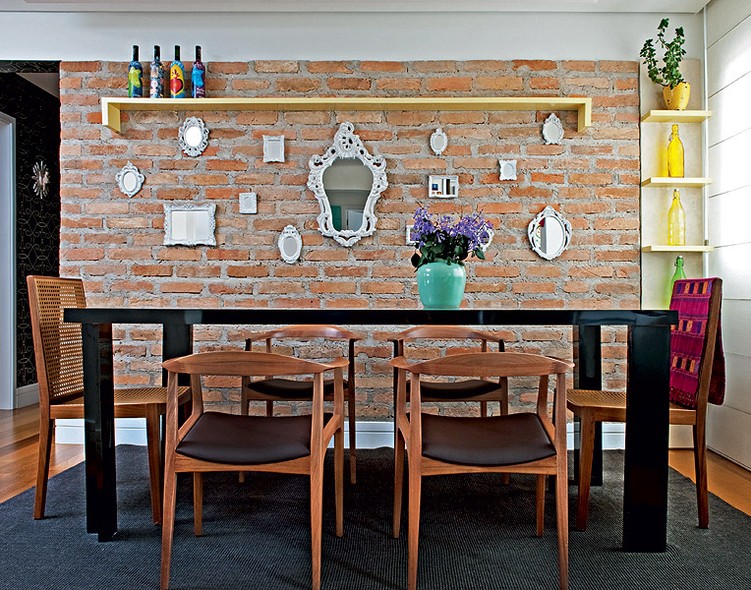 Na sala de jantar, criada pela designer de interiores Amanda Borges, a parede de tijolos é valorizada por vários espelhos, de diferentes tamanhos. O toque de cor fica por conta das garrafas decorativas, o vaso com flores e a manta sobre uma das cadeiras