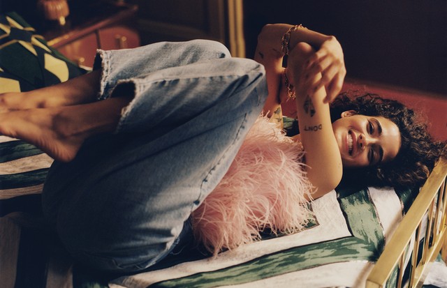 Moda basada en AtticoLa modelo posa tumbada en la cama sonriendo top rosacon plumas de avestruz, vaqueros Cavalier de tiro alto y pulseras bañadas en oro, todo de Attico (Foto: Luca Capri)