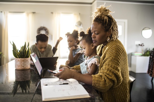 Mother working from home while holding toddler, family in background (Foto: Getty Images)