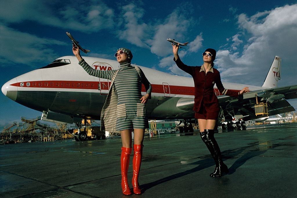 Two models posing on a tarmac in front of a Boeing 747 for TWA wearing, from left: a blue/ beige striped cotton dress with matching sweater and cap by Anna Modeller, enamel-framed sunglasses by Tropic-Cal and over-the-knee red patent boots by Latinas; rig (Foto: Conde Nast via Getty Images)