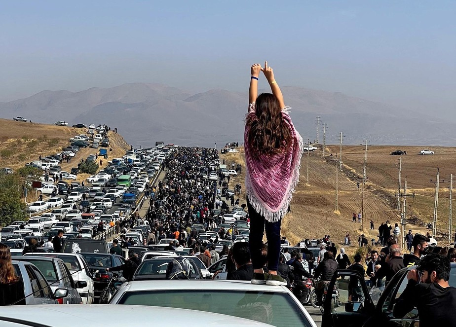 Mulher sem véu em cima de carro onde milhares de pessoas se dirigem para protesto na cidade iraniana onde Mahsa Amini está enterrada