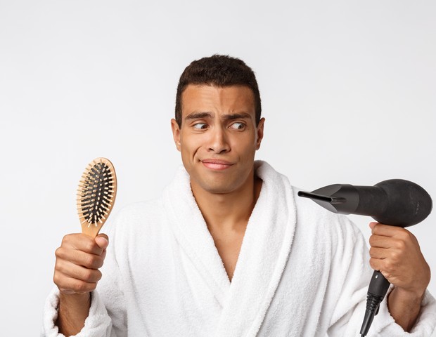 Portrait of mature funny beautiful tan-skinned african guy with curly hair in casual white bathrobe having problems with combing hair and hair dryer. (Foto: Getty Images/iStockphoto)