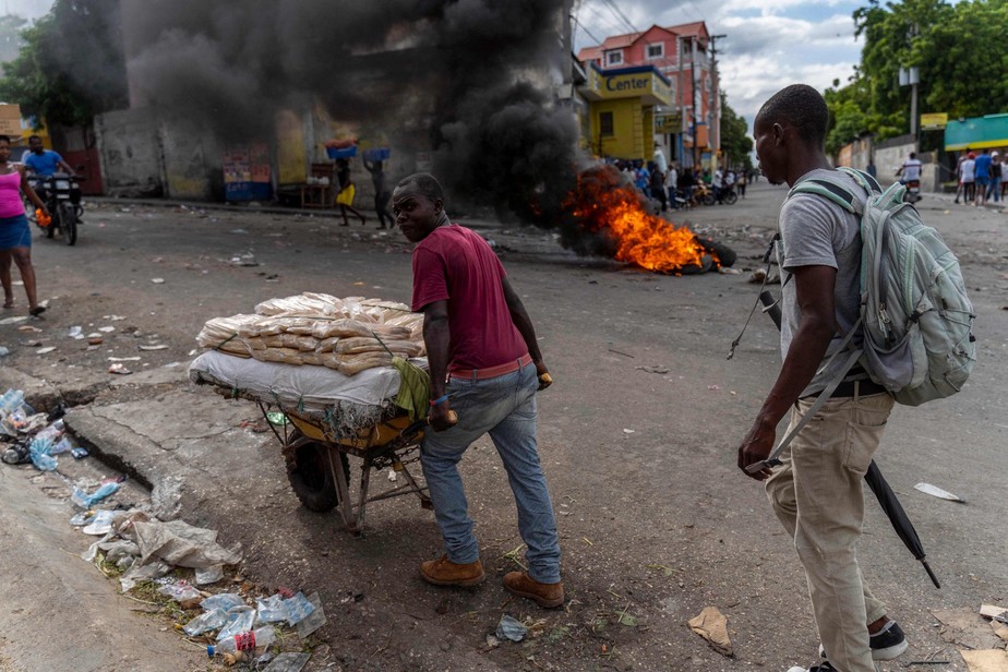 Um homem carrega um carrinho de mão em frente a pneus em chamas durante um protesto contra o primeiro-ministro haitiano Ariel Henry