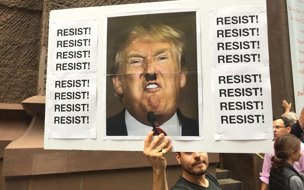 Manifestante exibe cartaz com Trump usando um bigode semelhante ao de Hitler em frente à Trump Tower, em Nova York, na segunda (14) (Foto: Orlando Moreira/TV Globo)