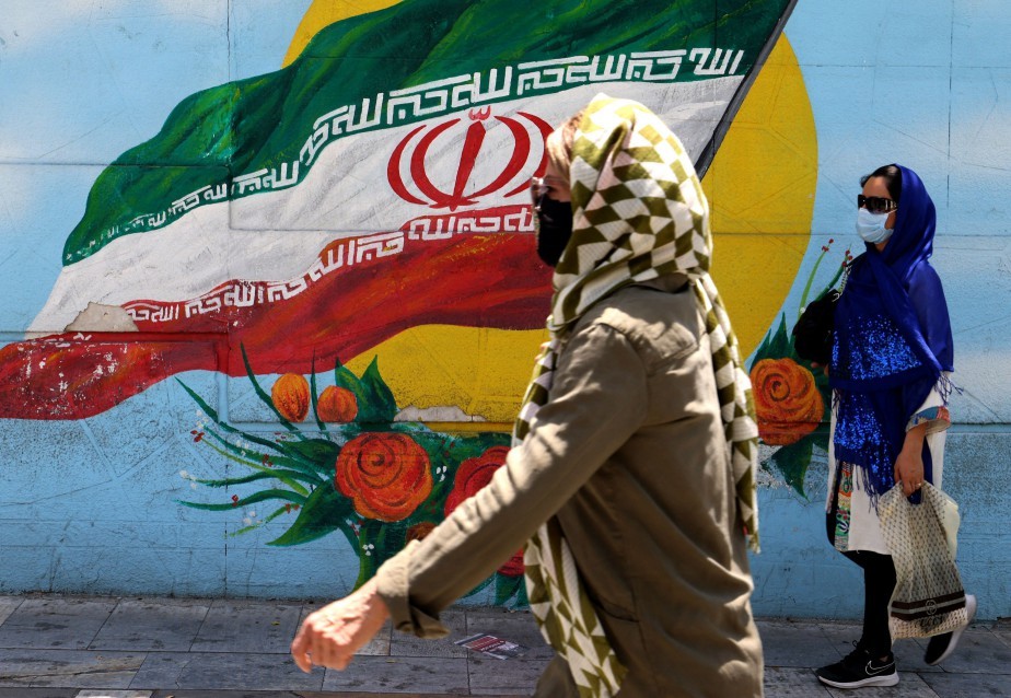 De máscara, mulheres caminham em frente à mural com a bandeira iraniana, em Teerã