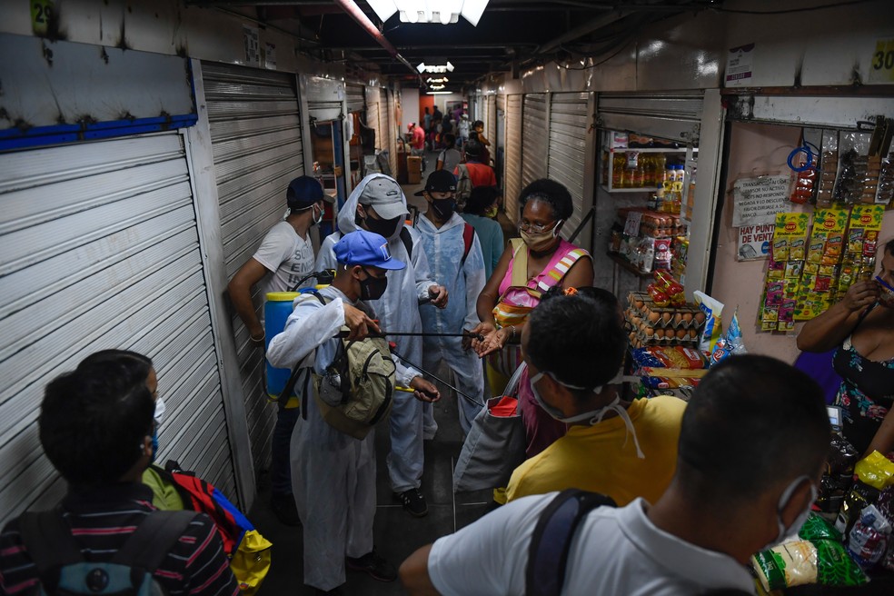 Um funcionário municipal com um traje de proteção pulveriza desinfetante nas mãos de um cliente em um mercado no bairro de Petare em Caracas, na Venezuela — Foto: Federico Parra/AFP