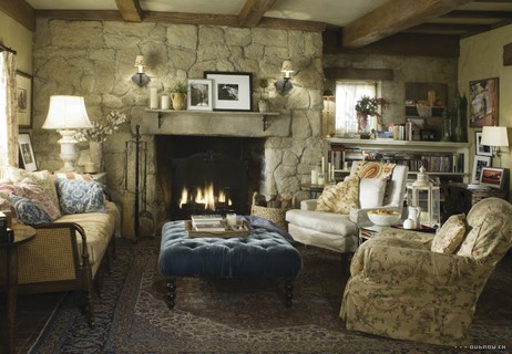Na sala de estar. uma lareira inserida em parede de pedra contrasta com o luxuoso pufe de veludo azul