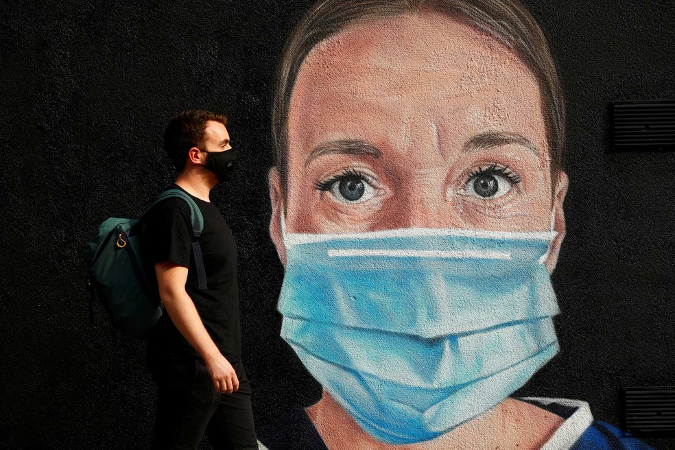 Um homem usando uma máscara passa por um mural de um trabalhador médico no centro da cidade de Manchester, na Inglaterra — Foto: Jason Cairnduff/Reuters