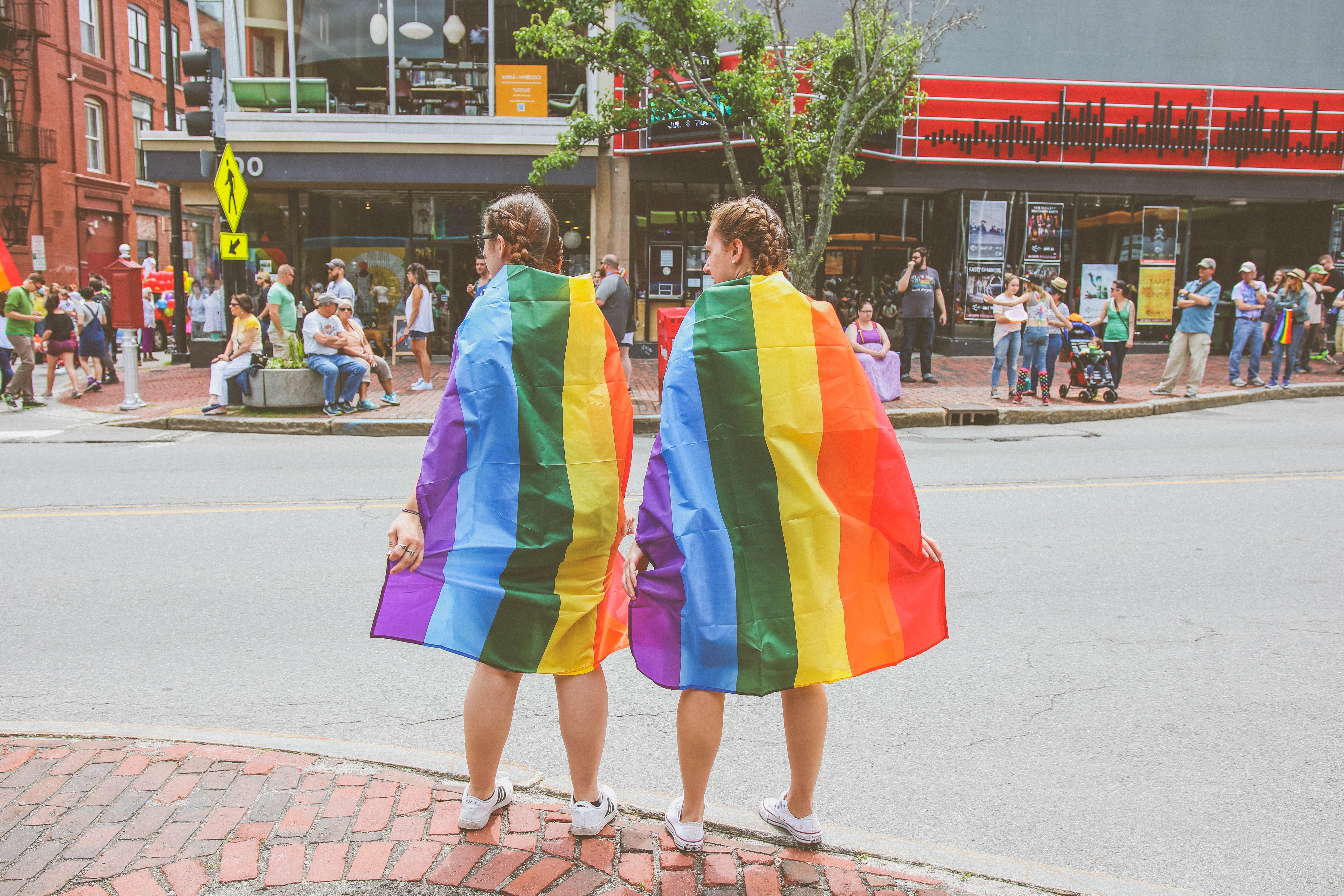 Você sabe o que significam as letras de LGBTQI+? Acima: mulheres de costas paradas em um esquina com a bandeira da comunidade LGBTQI+ amarrada ao pescoço (Foto: Mercedes Mehling/Unsplash)