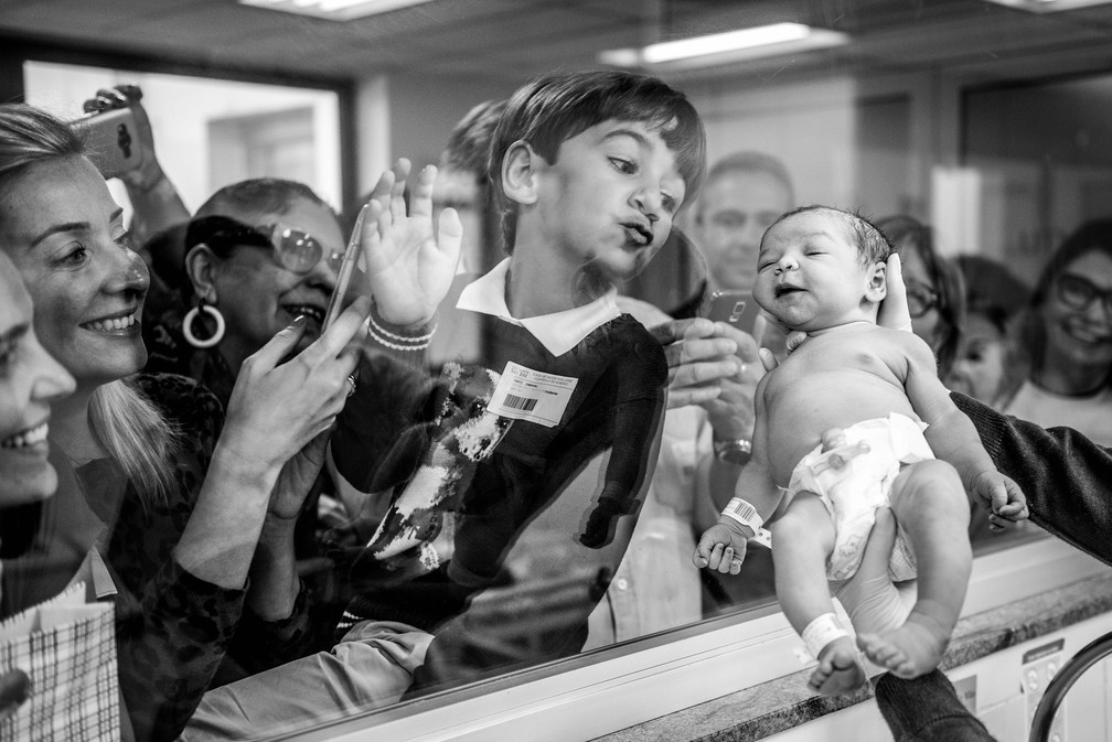 Irmão manda beijo pelo vidro para a irmãzinha recém-nascida em imagem premiada pela Associação Internacional de Fotógrafos Profissionais de Nascimento. (Foto: Daniela Justus/ Arquivo pessoal)