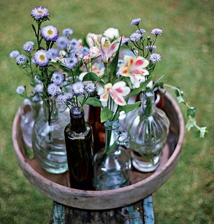 Nada de furos, fios ou quebra-quebra. Para montar este centro de mesa, basta reunir garrafas de diferentes cores e formatos e organizá-las em uma bandeja de madeira. Colha algumas flores no jardim e pronto!