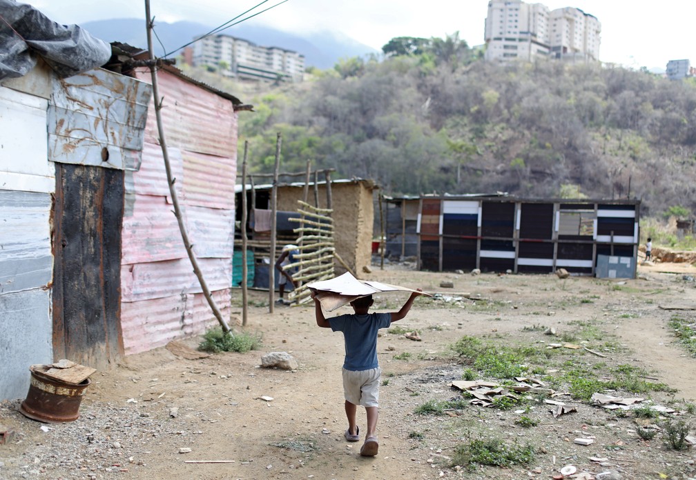 Um menino carrega um pedaço de alumínio perto de pequenas casas feitas de barro, paus e folhas de alumínio criadas em um terreno baldio e onde famílias estão se instalando por não podem mais pagar aluguel no município de Sucre, perto de Caracas, na Venezuela, em junho de 2020 — Foto: Manaure Quintero/Reuters